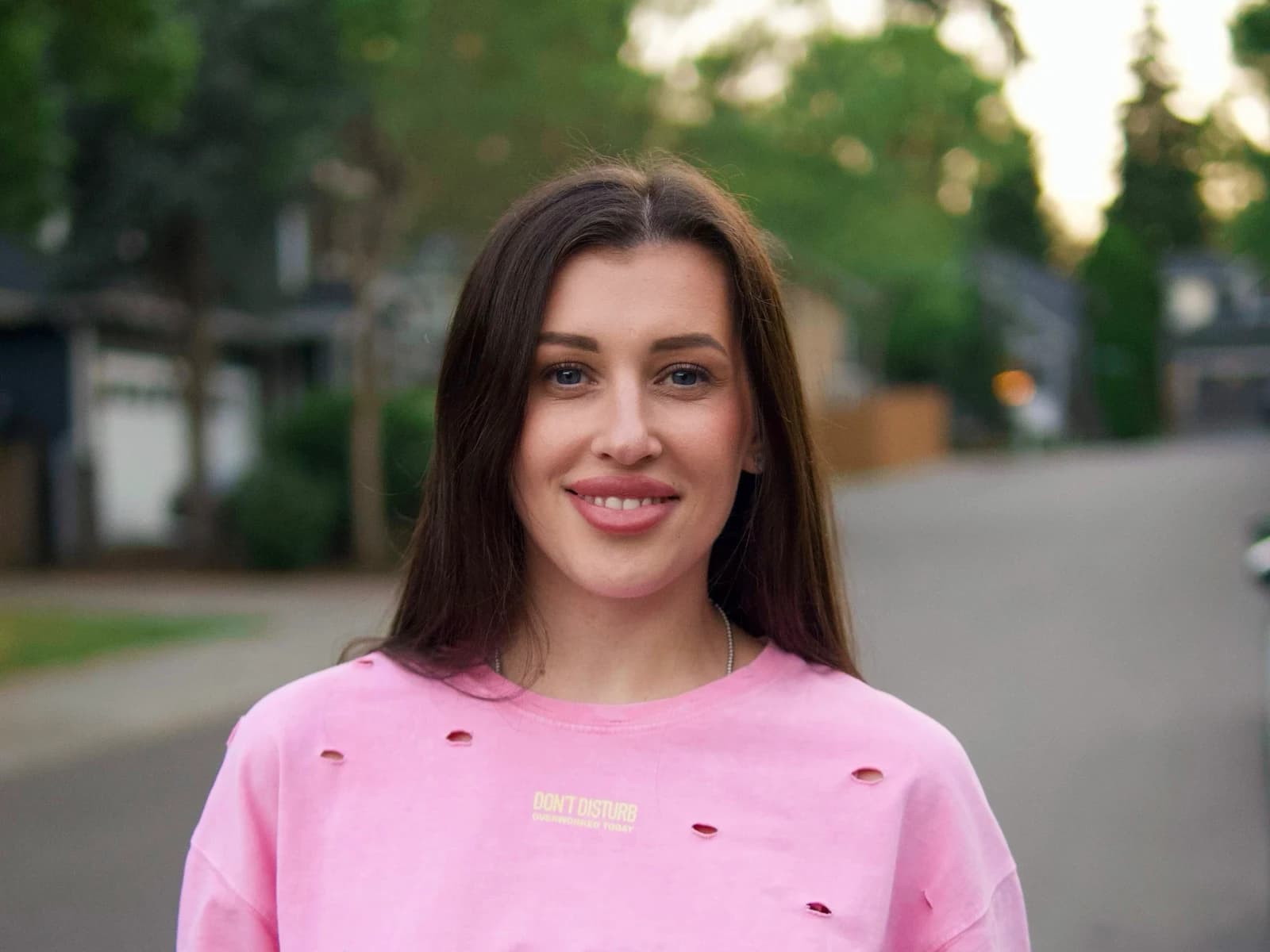 A portrait of a smiling woman with long dark hair wearing a distressed pink t-shirt, against a blurred street background. A quote is overlaid on the image in white font on a black textured background: "Great color isn't applied it's built, one controlled layer at a time."