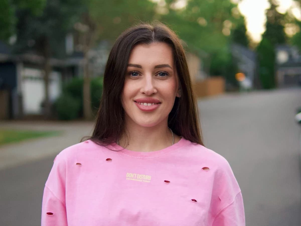 A portrait of a smiling woman with long dark hair wearing a distressed pink t-shirt, against a blurred street background. A quote is overlaid on the image in white font on a black textured background: "Great color isn't applied it's built, one controlled layer at a time."